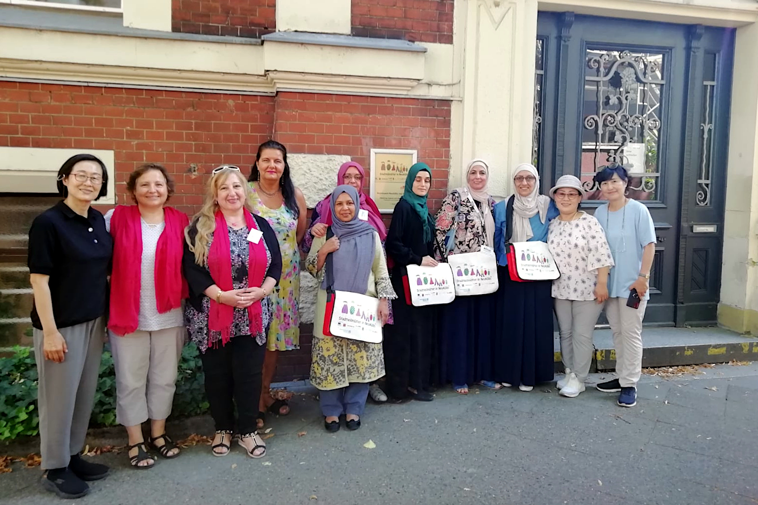 Elf Frauen stehen für ein Gruppenfoto vor einem Gebäude. Manche tragen ein Kopftuch und Stadtteilmütter-Taschen.