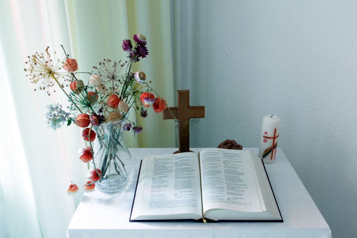 Auf einem Tisch liegt eine offene Bibel, es stehen Blumen, ein Kreuz und eine Kerze daneben.